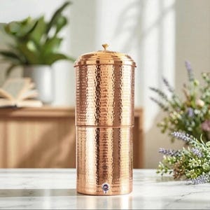 May include: A cylindrical copper water dispenser featuring a hammered texture throughout its exterior surface. The vessel includes a removable lid with a small knob handle at the top and a circular spigot located near the base for dispensing liquid.