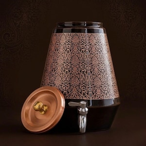 Puede incluir: Un dispensador de agua de color marrón oscuro y forma cónica con un intrincado patrón de damasco repetitivo en color cobre. El recipiente incluye un grifo de metal plateado en la base y una tapa circular de color cobre con un pomo dorado.