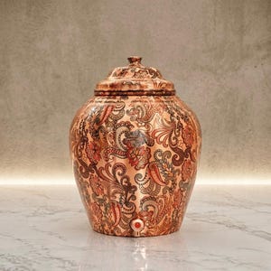 May include: A copper water dispenser with a rounded body and a matching lid. The surface is covered in an intricate, repeating floral and paisley pattern in shades of brown, orange, and grey. A small red circular tap is located at the base.