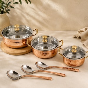 May include: A set of three hammered copper pots with stainless steel interiors, glass lids, and gold-toned handles, accompanied by three matching hammered copper spoons with stainless steel bowls, arranged in a graduated size display.