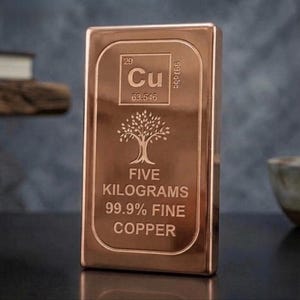 May include: A rectangular copper bar with a polished finish. The front features the periodic table symbol Cu, the atomic number 29, and the atomic weight 63.546. Below is a tree icon, the text FIVE KILOGRAMS, and 99.9% FINE COPPER.