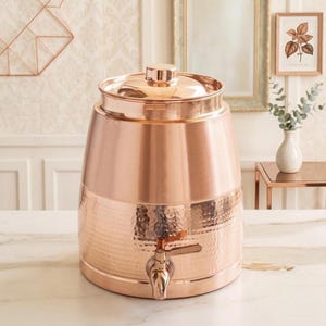 May include: A cylindrical copper water dispenser featuring a polished finish on the upper section and a hammered texture on the lower half. It includes a matching lid and a metal spigot attached to the front for dispensing liquid contents.