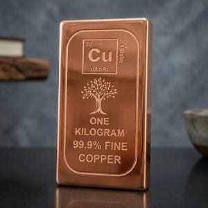 May include: A rectangular copper bar featuring the periodic table symbol Cu, atomic number 29, and atomic weight 63.546. The bar is engraved with a tree icon, the text ONE KILOGRAM, 99.9% FINE, and COPPER. The surface has a polished metallic finish.