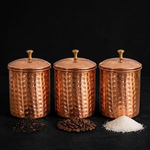 Puede incluir: Tres recipientes cilíndricos de cobre con textura martillada y tapas a juego rematadas con pequeños pomos de color latón. Cada recipiente contiene una sustancia diferente: té a granel, granos de café tostados y azúcar blanca granulada.