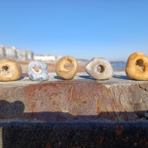 May include: Five naturally formed stones with holes, known as hag stones, are arranged in a row. The stones vary in color from light tan to grey and feature smooth, rounded surfaces with distinct, naturally occurring perforations through the center.