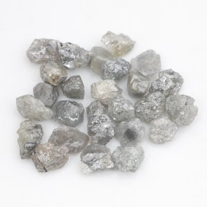 Peut inclure: Une collection de petits diamants bruts et non taillés dans diverses nuances de gris, d'argent et de blanc translucide. Chaque pierre présente une texture de surface dentelée, irrégulière et cristalline, révélant des facettes naturelles.