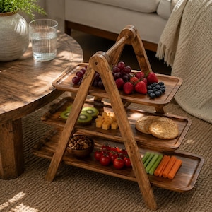 May include: A three-tiered wooden serving stand with an A-frame design. The top tray holds grapes, strawberries, and blueberries. The middle tray features kiwi slices, cheese cubes, and round crackers. The bottom tray contains nuts, cherry tomatoes, celery, and carrot sticks.