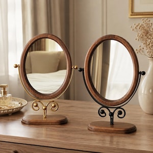 May include: Two oval tabletop mirrors with wooden frames and wooden bases. One mirror features a gold-toned metal stand with scrollwork details, while the other mirror features a black-toned metal stand with matching scrollwork accents.