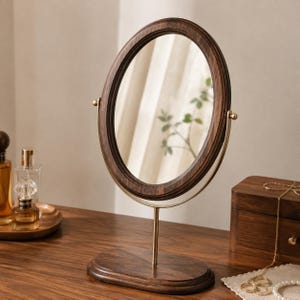 May include: An oval tabletop mirror featuring a dark brown wood frame and a gold-toned metal stand. The mirror is mounted on a matching dark brown wooden oval base, allowing for rotation. The design includes a slim metal support arm.