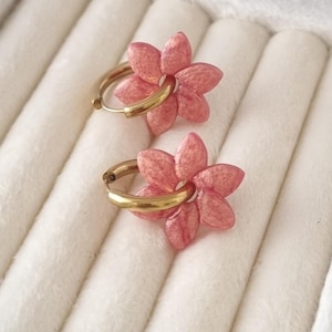Peut inclure: Une paire de boucles d'oreilles créoles avec une breloque en forme de fleur rose à cinq pétales, dotée d'une finition texturée et scintillante. Les anneaux sont en métal doré lisse et poli, chaque fleur est fixée à l'anneau circulaire.