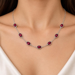 Peut inclure: Un collier à chaîne argentée orné de six pierres précieuses rouge foncé, de forme ovale et à facettes, réparties uniformément. Le collier est composé d'une fine chaîne à maillons reliant chaque sertissage, créant un design symétrique.