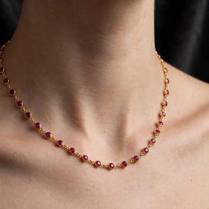 Puede incluir: Un collar de cadena dorada que presenta una serie de piedras preciosas de color rojo intenso, redondas y engastadas en bisel, espaciadas uniformemente. El collar consta de finos eslabones dorados que conectan cada engaste individual.