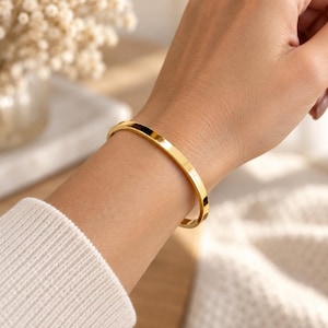 Könnte beinhalten: Ein polierter Armreif aus goldfarbenem Metall mit einer glatten, flachen Oberfläche und einer kreisförmigen Form. Das Armband zeichnet sich durch ein schlichtes, durchgehendes Banddesign aus, das bequem am Handgelenk anliegt.