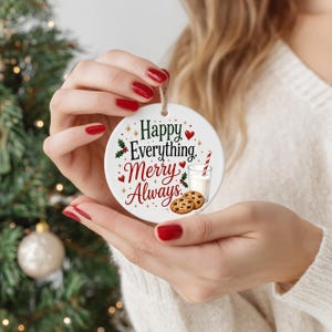 Op de afbeelding: Een ronde witte keramische hanger met de tekst Happy Everything Merry Always in zwarte en rode letters. Het ontwerp bevat illustraties van twee koekjes met chocoladestukjes, een glas melk met een rood-wit gestreept rietje en hulst.