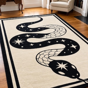 Può includere: Un tappeto rettangolare con sfondo color crema e un bordo nero spesso. Il centro presenta un grande motivo a serpente nero sinuoso decorato con forme di stelle bianche e un dettagliato motivo a scaglie su parti del corpo.