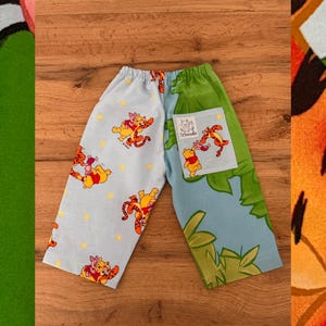 May include: Pants featuring a split design with a light blue side patterned with Winnie the Pooh, Tigger, and Piglet characters alongside yellow stars, and a green side with a patch pocket displaying the text Wrecubs and cartoon characters.