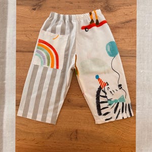 May include: Trousers featuring an asymmetrical design with one leg in grey and white vertical stripes and the other in white with colourful illustrations of a zebra wearing a party hat, a blue balloon, and a skateboard. A pocket displays a rainbow.