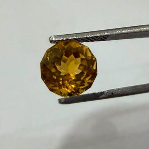 Könnte beinhalten: Ein facettierter, runder, goldgelber Edelstein, der von einer silberfarbenen Metallpinzette gehalten wird. Der Stein weist einen geometrischen Schliff mit vielen Facetten auf, der das Licht reflektiert und für ein funkelndes Aussehen sorgt.