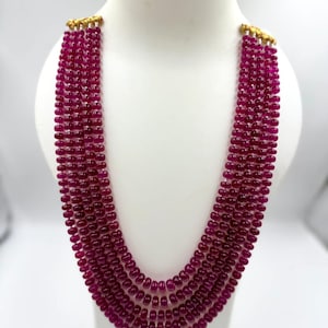 Peut inclure: Un collier multi-rangs composé de cinq rangées de perles rondes et lisses de couleur magenta profond. Les brins sont réunis en haut par des éléments en métal doré, créant un effet superposé en forme de U sur un présentoir blanc.