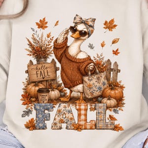 Puede incluir: Una sudadera de cuello redondo color crema con un gráfico de un pato que lleva un suéter marrón, gafas de sol y una diadema estampada. El pato sostiene un bolso floral junto a un letrero de madera que dice hello FALL y varias calabazas.