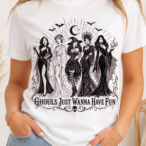 A person wearing a white t-shirt featuring a black graphic of five witches and the text Ghouls Just Wanna Have Fun.
