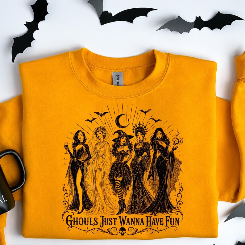 A mustard yellow crewneck sweatshirt laid flat on a white background with a black gothic witch design and paper bats.
