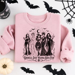 A light pink crewneck sweatshirt centered on a white backdrop, printed with a black gothic witch design and surrounded by bats and a pumpkin.
