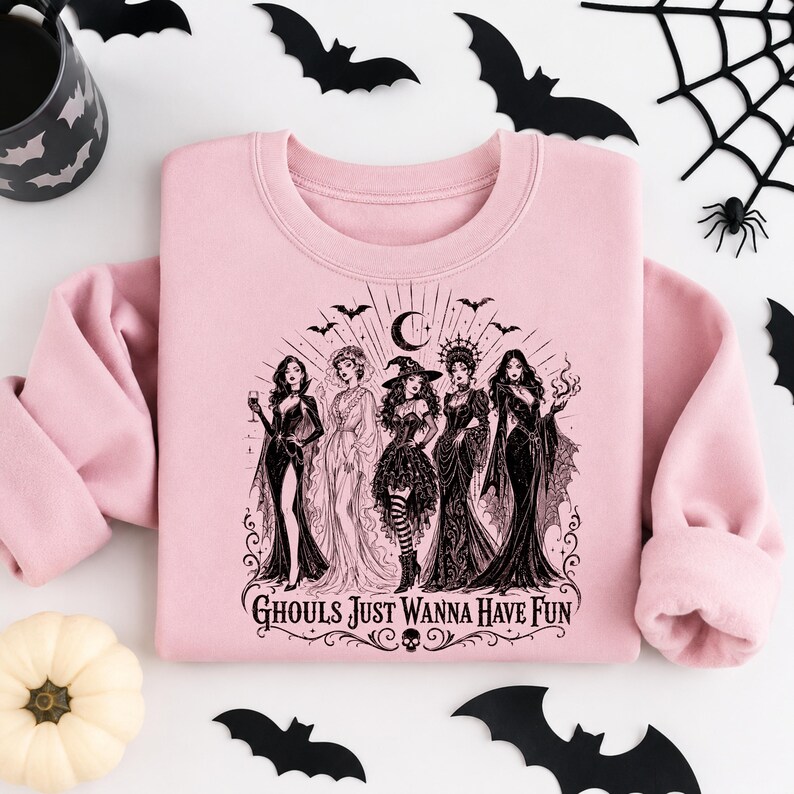 A light pink crewneck sweatshirt centered on a white backdrop, printed with a black gothic witch design and surrounded by bats and a pumpkin.