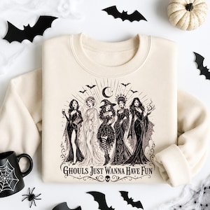 A cream sweatshirt with a black Ghouls Just Wanna Have Fun witch design, surrounded by a white pumpkin, bats, and a spider mug.