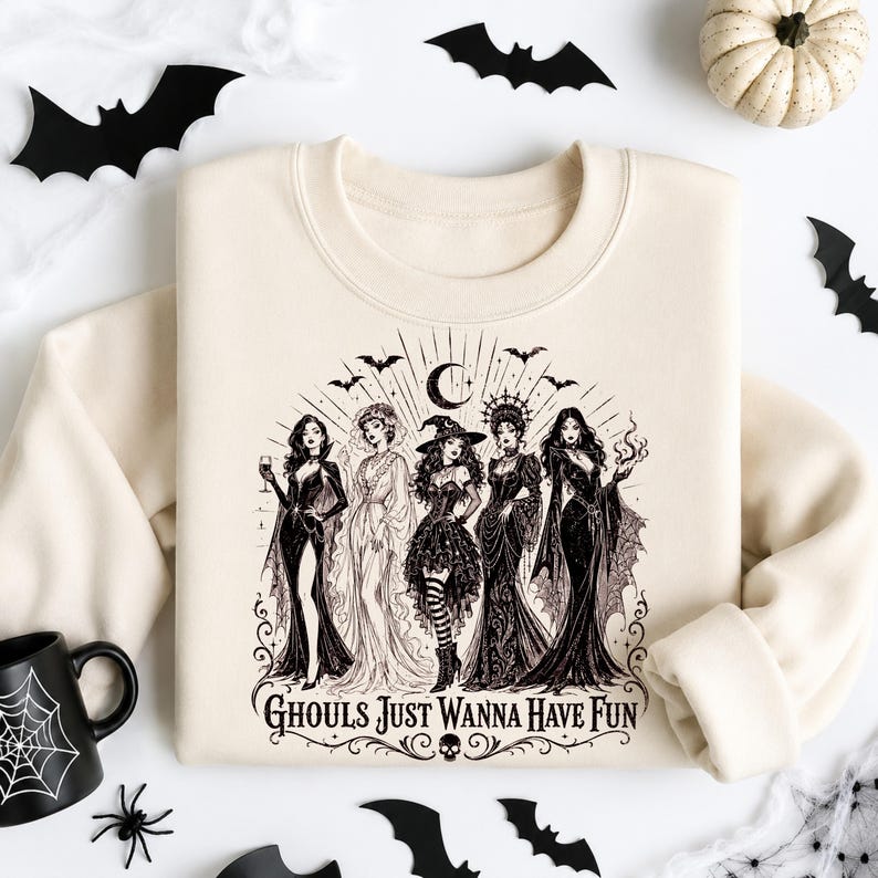 A cream sweatshirt with a black Ghouls Just Wanna Have Fun witch design, surrounded by a white pumpkin, bats, and a spider mug.