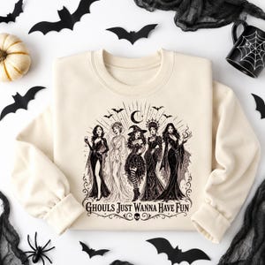 A cream-colored crewneck sweatshirt centered on a white background with a black witch coven graphic and Halloween props.