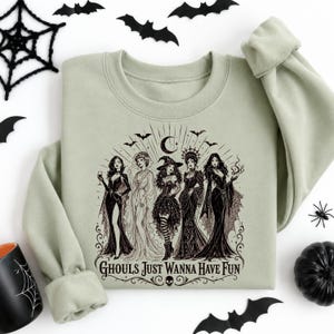 A sage green crewneck sweatshirt laid on a white surface with a black witch illustration and various spooky decorative elements.