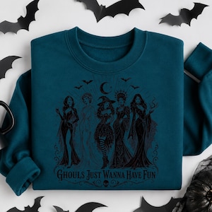 A dark teal crewneck sweatshirt displayed with a black witch coven graphic, paper bats, and a black pumpkin.