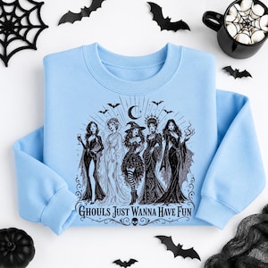 A light blue crewneck sweatshirt featuring a black graphic of witches and bats on a white background with a coffee mug and spiderweb.