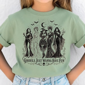 A person wearing a sage green t-shirt printed with a black and white illustration of five witches and the text Ghouls Just Wanna Have Fun.