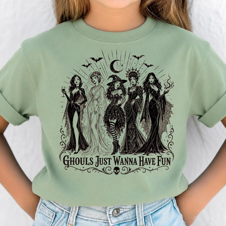 A person wearing a sage green t-shirt printed with a black and white illustration of five witches and the text Ghouls Just Wanna Have Fun.
