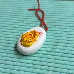 May include: A handmade pendant with a white oval base, decorated with a yellow and red design, reminiscent of a deviled egg. It has a red string for hanging. The pendant is set against a light teal background.