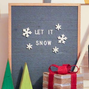 Puede incluir: Un tablero de letras gris con la frase "Let it snow" escrita en letras blancas. Hay decoraciones de copos de nieve blancos alrededor de la frase. Un regalo envuelto con una cinta roja y un &aacute;rbol de Navidad de madera verde y amarillo est&aacute;n en primer plano.