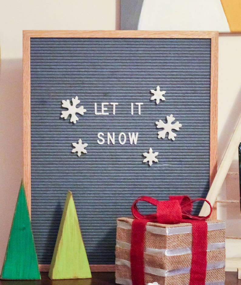 Puede incluir: Un tablero de letras gris con la frase "Let it snow" escrita en letras blancas. Hay decoraciones de copos de nieve blancos alrededor de la frase. Un regalo envuelto con una cinta roja y un &aacute;rbol de Navidad de madera verde y amarillo est&aacute;n en primer plano.