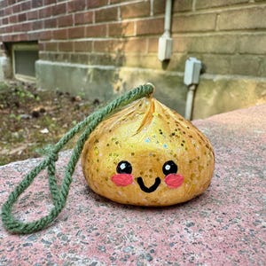 May include: A round, gold-colored ornament with a glittery finish, featuring a hand-painted face with black eyes, a black smile, and pink cheeks. A thick, green yarn loop is attached to the top for hanging, creating a decorative, rounded object.
