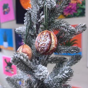 May include: Two round, golden-brown sourdough bread ornaments hanging from a flocked artificial pine tree. Each ornament features a detailed, scored crust with a dusting of white flour, suspended by a thin, dark green string tied to the branches.