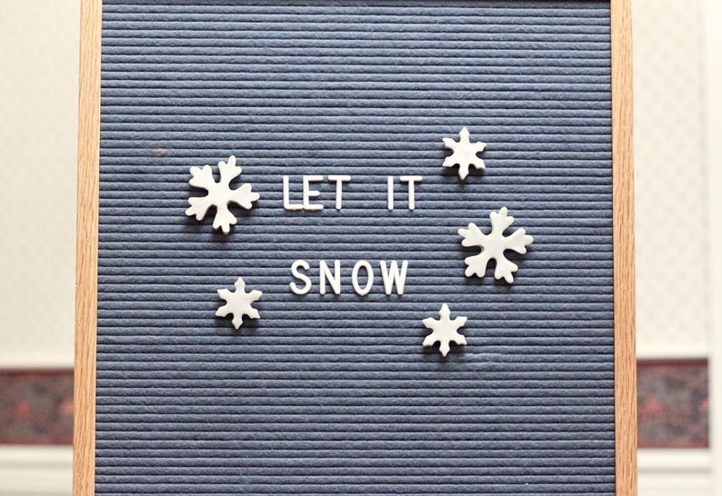 Puede incluir: Un tablero de letras de fieltro azul con letras blancas que deletrean "Let it snow" y decoraciones de copos de nieve blancos.