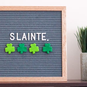 May include: A grey letter board with a wooden frame displays the word "SLAINTE."  Four green shamrock shapes are arranged below the word.