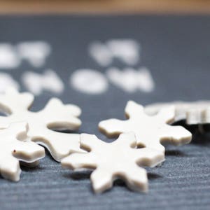Puede incluir: Adornos blancos con forma de copo de nieve sobre una superficie gris oscuro. Los adornos est&aacute;n hechos de un material blanco liso y tienen un dise&ntilde;o ligeramente en relieve. El texto "Let it snow" es visible en el fondo.