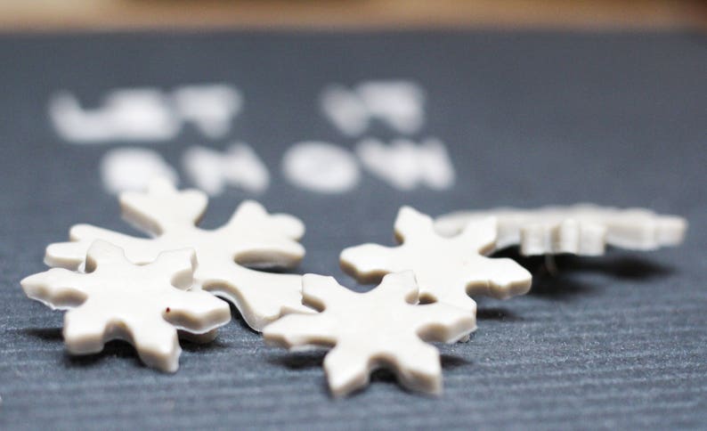 Puede incluir: Adornos blancos con forma de copo de nieve sobre una superficie gris oscuro. Los adornos est&aacute;n hechos de un material blanco liso y tienen un dise&ntilde;o ligeramente en relieve. El texto "Let it snow" es visible en el fondo.