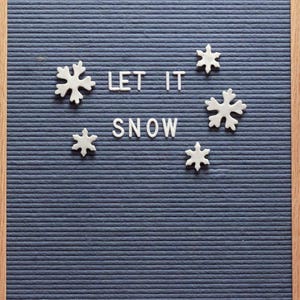 Puede incluir: Un tablero de letras de fieltro azul con un marco de madera. El tablero tiene letras blancas que deletrean "LET IT SNOW" y formas de copos de nieve blancas.