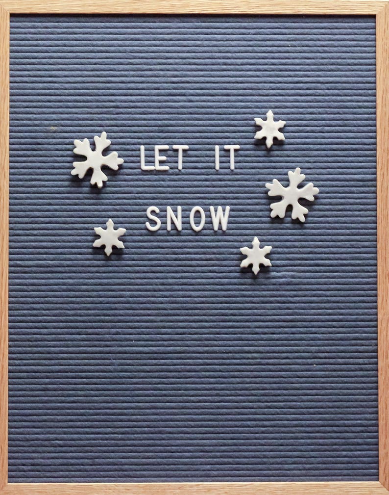 Puede incluir: Un tablero de letras de fieltro azul con un marco de madera. El tablero tiene letras blancas que deletrean "LET IT SNOW" y formas de copos de nieve blancas.