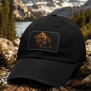 May include: A black baseball cap featuring a rectangular patch on the front panel. The patch displays a detailed illustration of a brown bison in a charging stance. The cap has a curved brim and stitched eyelets on the crown for ventilation.
