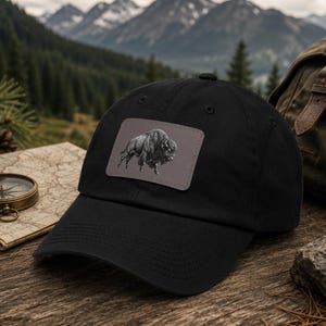 May include: A black baseball cap featuring a rectangular patch on the front panel. The patch displays a detailed, monochromatic illustration of a bison in profile, rendered in shades of gray and black against a solid, muted gray background.