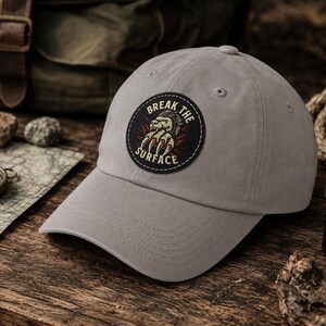 Pode incluir: Um boné de beisebol cinza-claro com um emblema circular preto na frente. O emblema apresenta um gráfico de uma mão de criatura com garras e o texto "BREAK THE SURFACE" arqueado em torno da ilustração em uma fonte de cor clara.